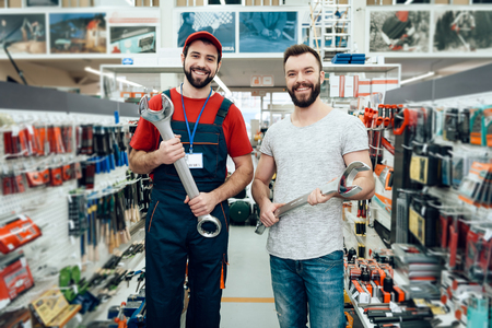 Salesman in red shirt and baseball cap and bearded client posing with giant wrenches in power tools store.の写真素材