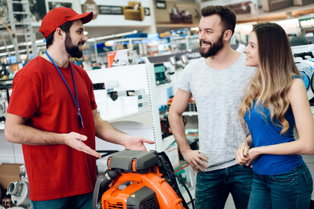 Salesman in red shirt and baseball cap is explaining details about leaf blower to couple of clients in power tools store.の写真素材