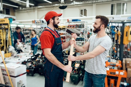 Salesman in red shirt and baseball cap is giving bearded client new giant hammer in power tools...