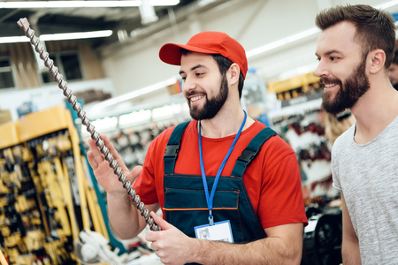 Salesman in red shirt and baseball cap is showing bearded client new giant drill in power tools...