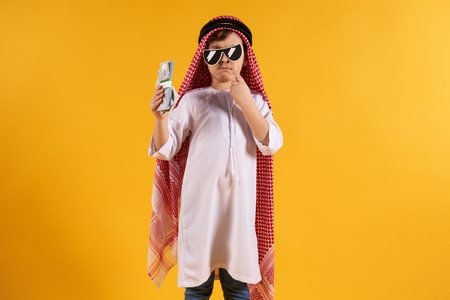 Arabian boy in keffiyeh holds bundle of dollars, hiding behind sham glasses. Thug life. Isolated on yellow background.の写真素材