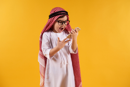 Arab smart boy in kufiya screams on smartphone. Isolated on yellow background. Studio portrait.の写真素材