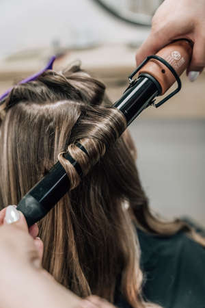 Female hands of a hair master. Close-up of blond hair on a curling iron. Curling curls. high quality photoの写真素材
