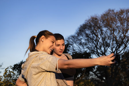 Two young women taking a selfie after working out in the park, conveying a sense of fun and friendship.Horizontal shot.Casual sport outfit.の写真素材