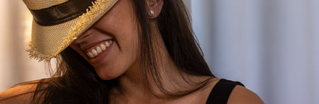 Young hispanic woman covering her eyes with a straw hat, shyly smiling and expressing joy, showing...