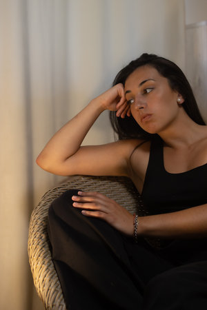 Young hispanic woman looking away, sitting in a wicker chair, contemplating her thoughts and...