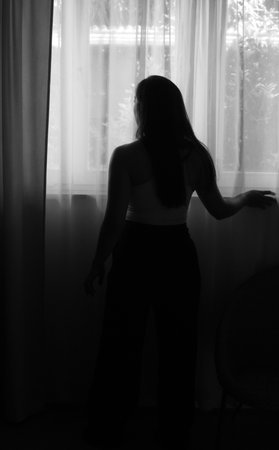 Pensive young woman standing in silhouette against a brightly lit window. Looking out with a thoughtful expression. Embodying concepts of contemplation. Solitude. And the quiet beauty of everyday lifeの写真素材