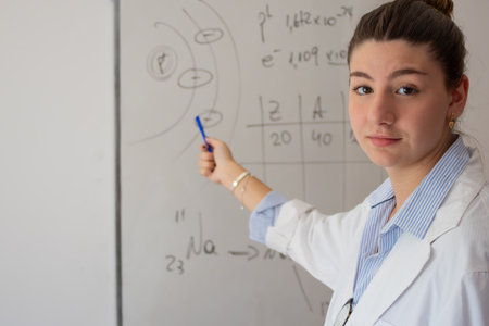 Young female teacher presenting a science lesson, pointing at a whiteboard with diagrams of atomic structure, chemical formulas, and mathematical calculations, showing chemistry teachingの写真素材