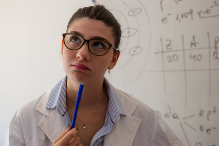 Young woman student in a lab coat and glasses, holding a pen to her chin while looking upwards, is deep in thought about scientific formulas and diagrams on a whiteboard in a classroom settingの写真素材