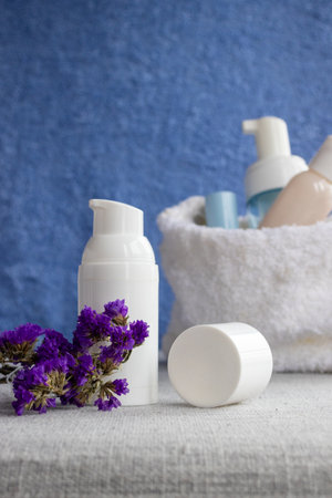 Skincare products for daily routine. Including lotion bottle with pump dispenser and cosmetic containers in white towel. Decorated with purple flowers against blue background. Promoting self careの写真素材