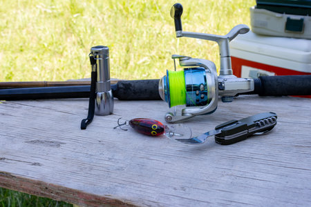 Fishing rod with reel, artificial lure, flashlight and multi tool laid on a rustic wooden table by green grass, evoking camping, fishing gear and relaxed outdoor adventureの写真素材