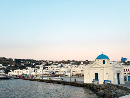 Mykonos island experiencing the sunset glow, presenting a whitewashed orthodox church with a blue dome by the harbor and traditional cycladic architecture along the coastの写真素材