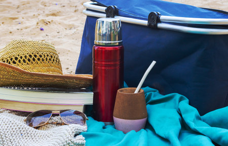 Beach or park picnic setup displaying a straw hat, books, sunglasses, a warmer, and a mate gourd on blue fabric near a cooler, representing leisurely outdoor relaxationの写真素材