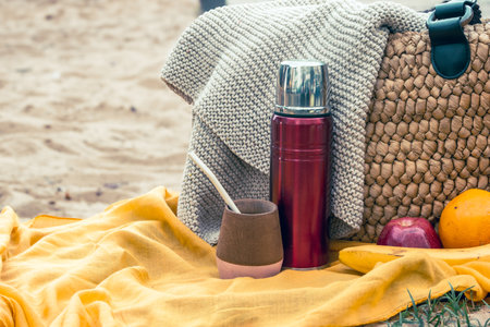 Picnic essentials on a yellow blanket in the sand, red thermos, mate gourd with , woven bag and assorted fruits sunny, healthy beach leisure sceneの写真素材