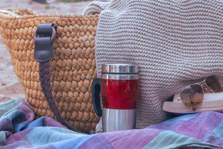 Picnic setup outdoors featuring a woven bag, knitted blanket, thermal mug, sunglasses, and a book resting on a striped blanket, evoking relaxation and leisure activitiesの写真素材