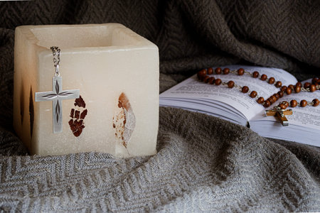 Christian devotional still life, rosary on a Bible and lit candle on textured gray fabric,...