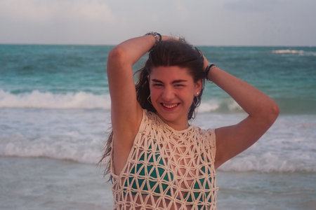 Young woman smiling with genuine happiness, hands in her hair as she stands waist-deep in turquoise ocean water on a carefree summer beach vacation, enjoying waves and sunshineの写真素材