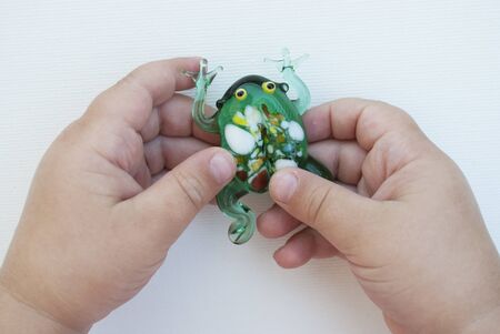 small green frog made of glass in the hands of a child on a white background. a child of three years is holding a toy. green frog made of glass with multicolored spotsの写真素材