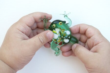 small green frog made of glass in the hands of a child on a white background. a child of three years is holding a toy. green frog made of glass with multicolored spotsの写真素材