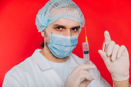 The doctor is holding a syringe and an ampoule with a vaccine. A young guy in a white coat, in a medical mask and gloves on a red background.の写真素材