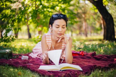 The girl lies on the grass and reads lectures in a notebook. A woman reads a book and listens to music in the park.の写真素材