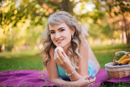 Beautiful girl on a picnic in the park eats a peach. Blonde lying on a purple bedspread resting.の写真素材
