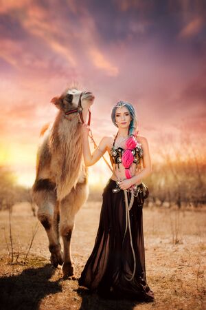 Beautiful girl with multi-colored braids in oriental clothes. Princess with a camel at sunset.の写真素材