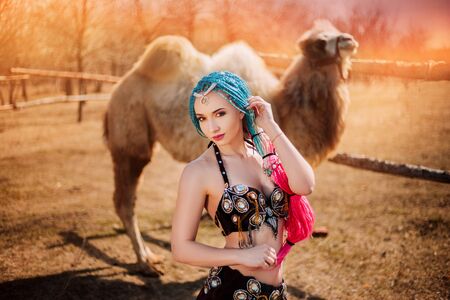 Beautiful girl with multi-colored braids in oriental clothes. Princess with a camel at sunset.の写真素材