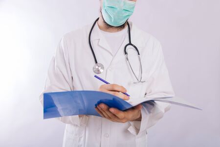 The doctor in a white coat in a medical mask holds in his hands a blue folder with a pen on a light background.の写真素材