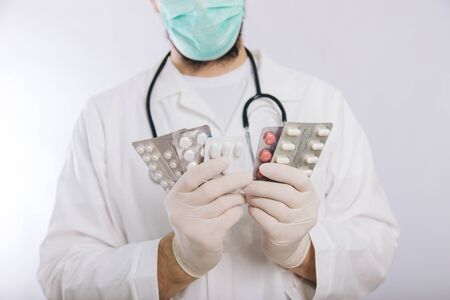 A doctor in a white coat, a medical mask and gloves holds pills. Types of drugs. Medical worker on a white background.の写真素材