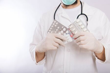 A doctor in a white coat, a medical mask and gloves holds pills. Types of drugs. Medical worker on a white background. Studio photoの写真素材