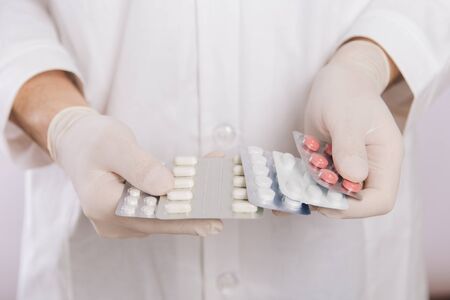A doctor in a white coat, a medical mask and gloves holds pills. Types of drugs. Medical worker on a white background. Studio photoの写真素材