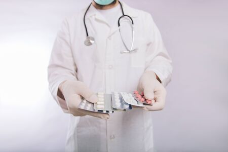 A doctor in a white coat, a medical mask and gloves holds pills. Types of drugs. Medical worker on a white background. Studio photoの写真素材
