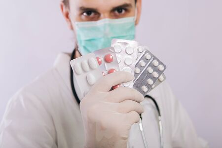 A doctor in a white coat, a medical mask and gloves holds pills. Types of drugs. Medical worker on a white background. Studio photoの写真素材