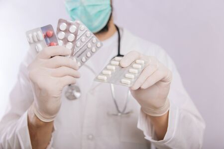 A doctor in a white coat, a medical mask and gloves holds pills. Types of drugs. Medical worker on a white background. Studio photoの写真素材