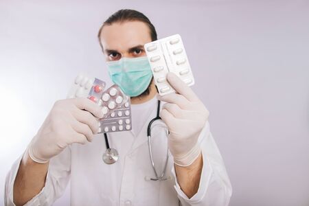A doctor in a white coat, a medical mask and gloves holds pills. Types of drugs. Medical worker on a white background. Studio photoの写真素材