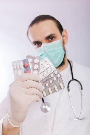 A doctor in a white coat, a medical mask and gloves holds pills. Types of drugs. Medical worker on a white background. Studio photoの写真素材