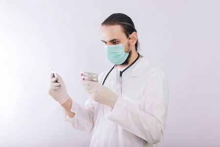 A doctor in a white coat, a medical mask and gloves holds pills. Types of drugs. Medical worker on a white background. Studio photoの写真素材