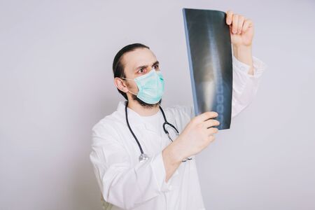 A doctor in a white coat and a medical mask holds in his hands pictures of lung X-ray on a white background. Examines X-ray pictures of a patientの写真素材