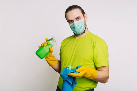 A guy in a green T-shirt and a medical mask holds in his hands a bulletzer with an antiseptic and a rag for dust on a white background. A man kills a coronavirus. disinfects the roomの写真素材