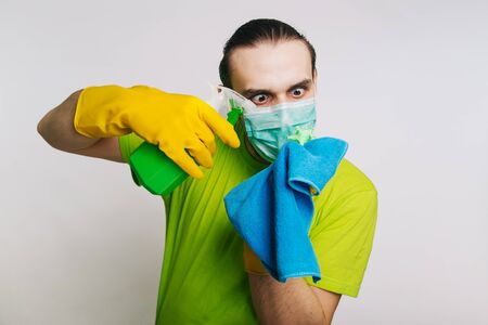A guy in a green T-shirt and a medical mask holds in his hands a bulletzer with an antiseptic and a rag for dust on a white background. A man kills a coronavirus. disinfects the roomの写真素材