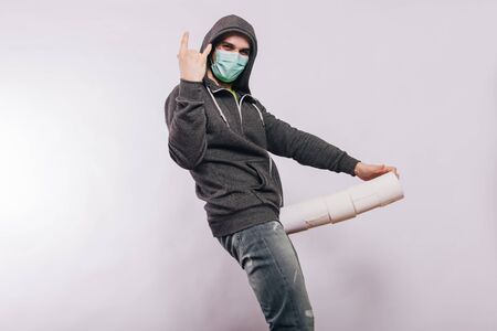 A guy in a gray sweatshirt on a white background in a medical mask holds a stack of toilet paper rolls in his groin.の写真素材