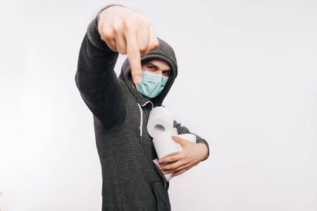 A guy in a gray sweatshirt on a white background in a medical mask holds rolls of toilet paper in his hands and shows an indefinite gesture of a fak....の写真素材