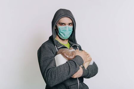 A guy in a gray sweatshirt on a light background in a medical mask holds essential products. A man in a panic buys toilet paper and buckwheat groats, rice.の写真素材