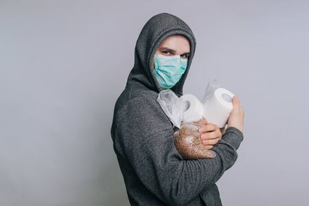 A guy in a gray sweatshirt on a light background in a medical mask holds essential products.の写真素材