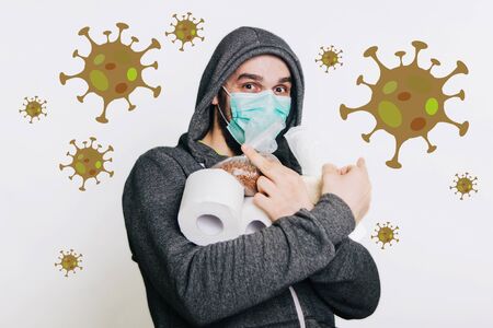 A guy in a gray sweatshirt on a light background in a medical mask holds his middle finger and shows a fac. A man in a panic buys toilet paper and buckwheat groats.の写真素材