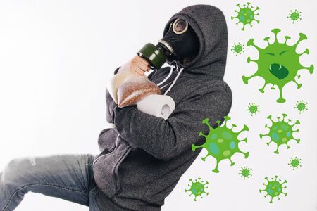 A guy in a gas mask and a gray hoody in a hood holds rolls of toilet paper and groats products in his hands on a white background. Running away from the Coronavirus . Panic during quarantine.の写真素材