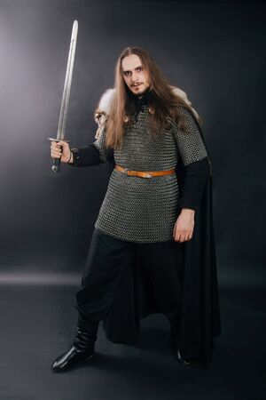 Warrior in armor with a sword. A guy with long hair and a beard, a fox collar. Dressed in chain mail, a black cloak and black pants holds a sword in his hands. Studio photo against a dark background.の写真素材