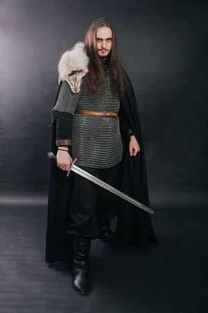 Warrior in armor with a sword. A guy with long hair and a beard, a fox collar. Dressed in chain mail, a black cloak and black pants holds a sword in his hands. Studio photo against a dark background.の写真素材