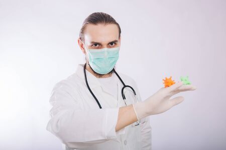 A doctor in a white coat and gloves on a white background holding a mock coronavirus in his hands. Shows a multi-colored virus.の写真素材
